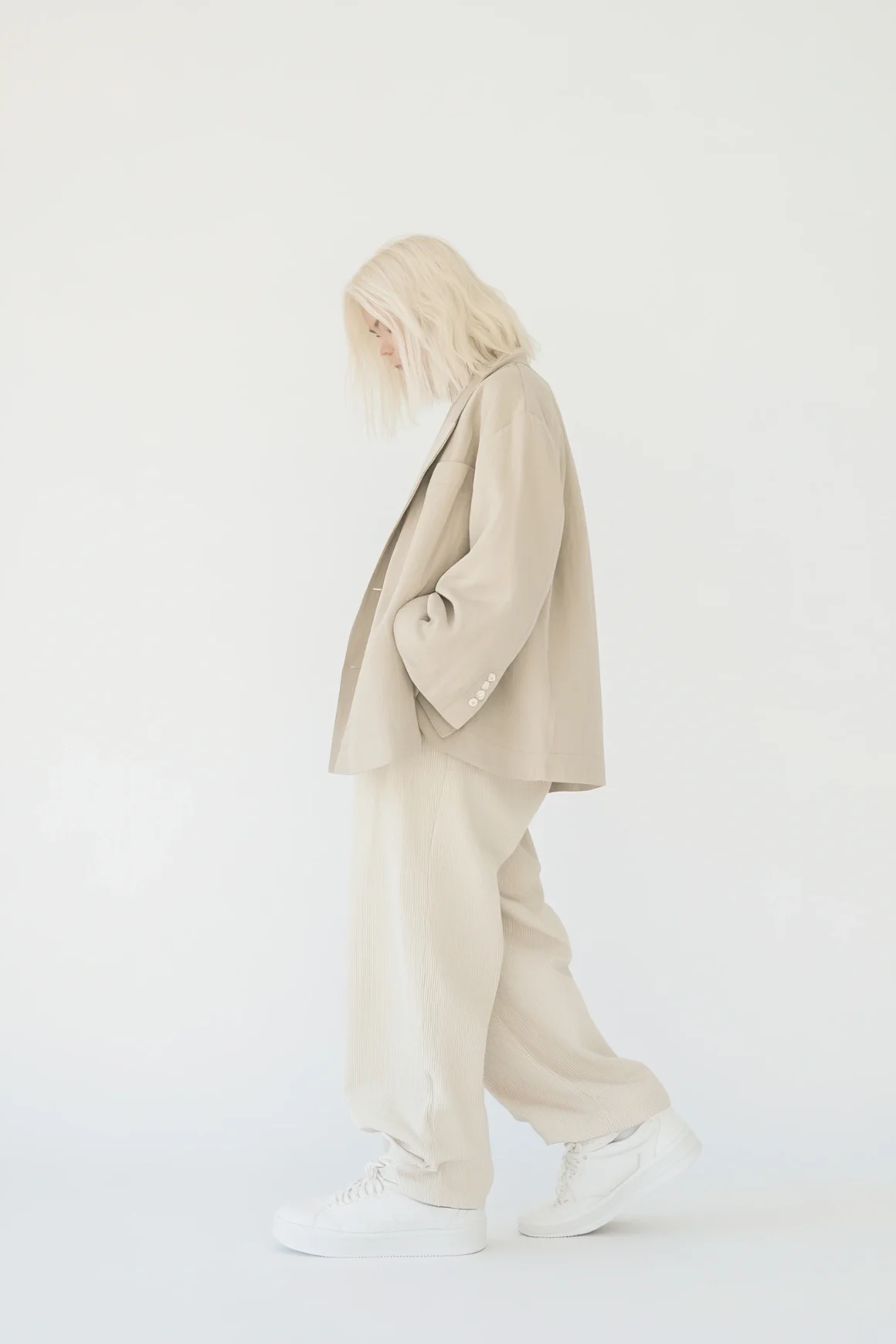 Woman in an oversized beige blazer and wide-leg trousers with white sneakers against a white backdrop.
