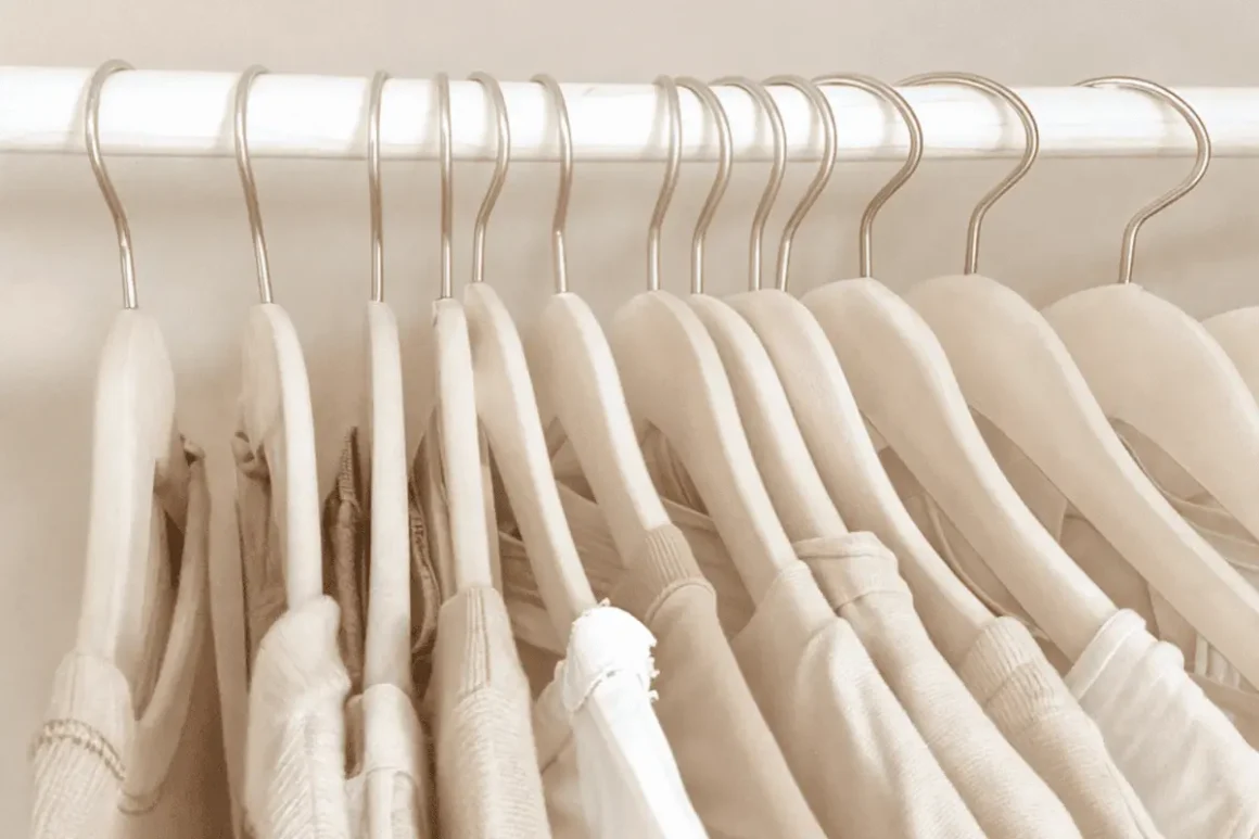 Row of neutrsl sweaters, shirts, and knitwear hanging on wooden hangers, a simple quiet luxury wardrobe in soft beige tones.