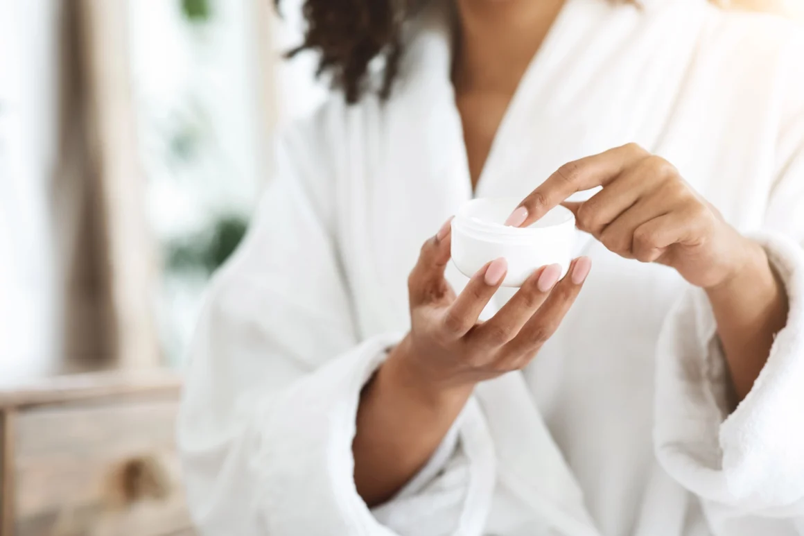 Woman in a white robe applying cream from a minimal white skincare jar.