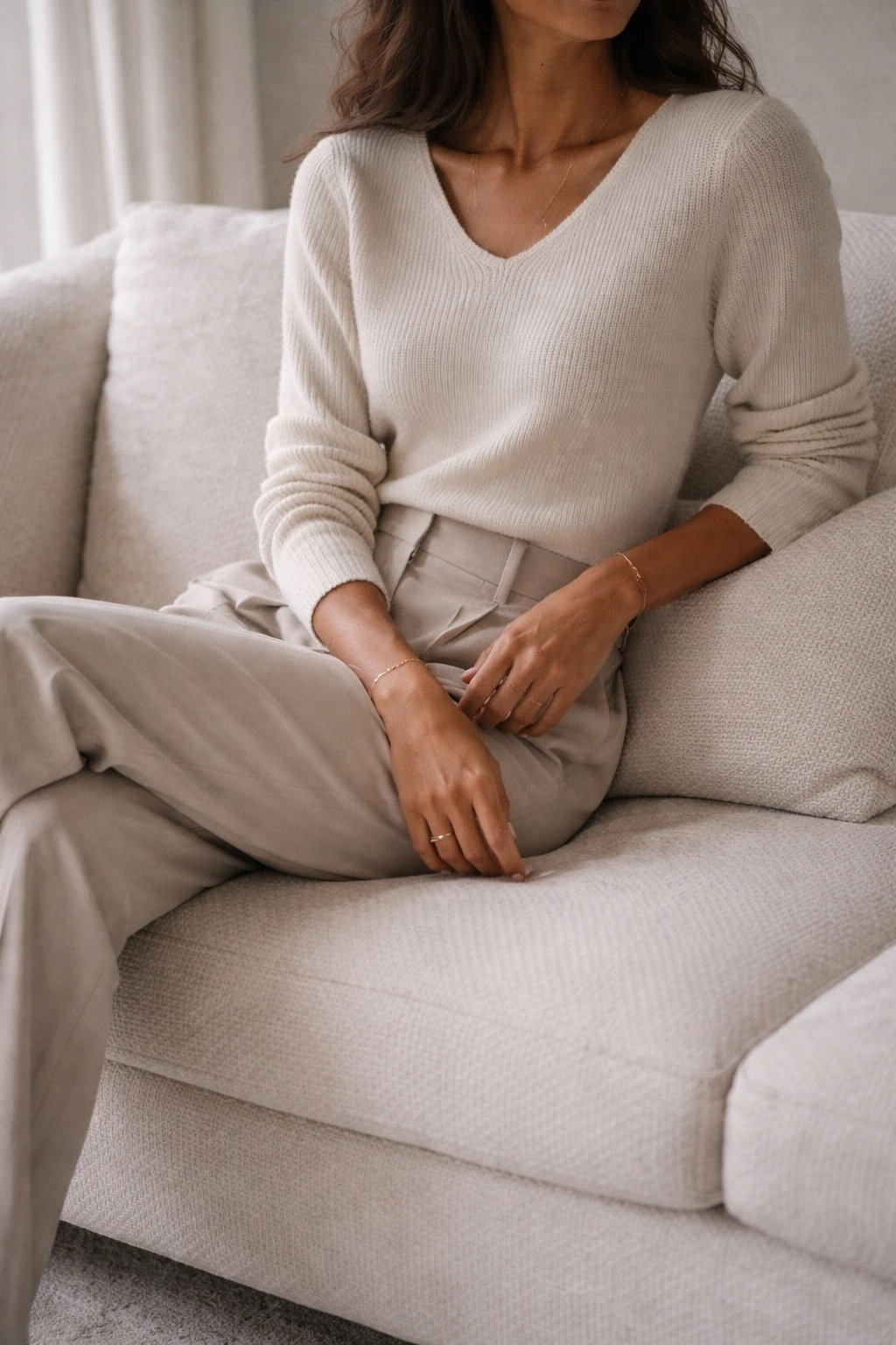 Woman seated in neutral armchair wearing a structured cream knit and tailored beige trousers, an example of comfortable but polished outfits.