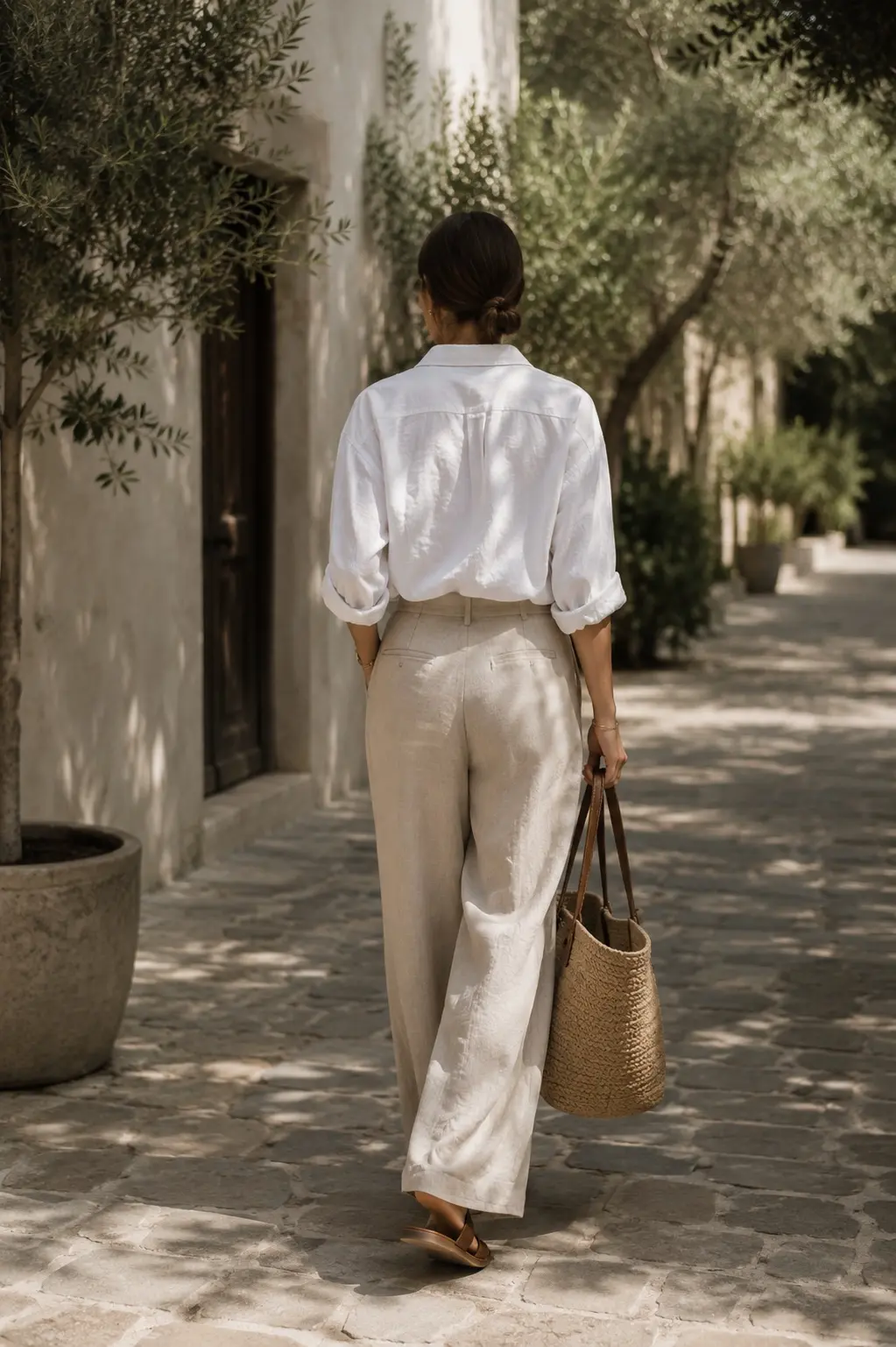 Back view of a woman walking beneath trees wearing the contents of her quiet luxury summer wardrobe: white shirt, linen trousers, summer tote, and comfortable sandals, that fade into the background as she enjoys a walk.